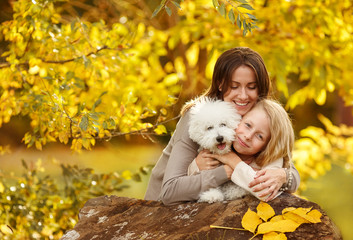 Happy family mom and daughter with little white dog hug and laugh sunny autumn day on the background of yellow leaves