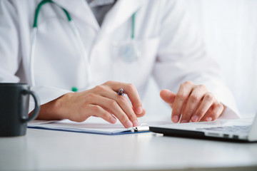 Male doctor working with laptop behind desk, writing on paperwork in a hospital.