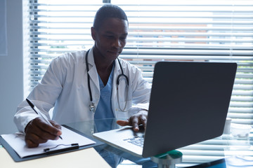Male doctor writing on a clipboard while using laptop at desk