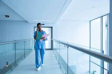 Female surgeon looking at medical reports while talking on mobile phone in corridor