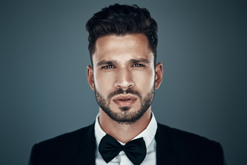 Charming young man in bow tie looking at camera while standing against grey background