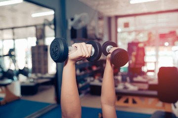 Hands are holding a black dumbbell in the gym with a backdrop of fitness room and sun light.