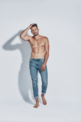 Full length of handsome young shirtless man looking at camera while standing against grey background