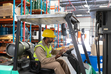 Male staff writing on clipboard while sitting on forklift in warehouse