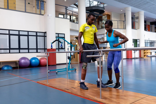 Female physiotherapist assisting disabled man walk with parallel bars in sports center