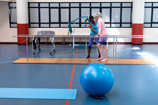 Female physiotherapist helping disabled senior woman walk with parallel bars in sports center