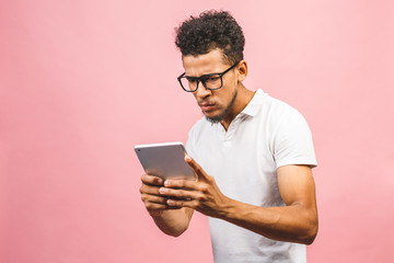 Excited african American male in glasses isolated on pink studio background hold tablet get good message, surprised black man receive get to know about sale offer or discount on pad.