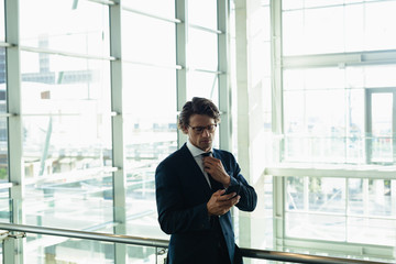 Businessman adjusting his tie while using mobile phone near railing in a modern office building