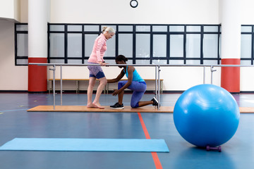 Female physiotherapist assisting disabled senior woman walk with parallel bars in sports center