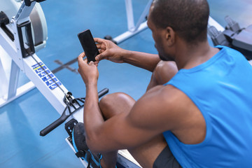 Fit man using mobile phone while exercising on rowing machine in fitness center
