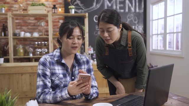 female barista chatting with girl guest in cafe store. asian chinese woman customer sitting at wooden table with laptop and showing waitress cellphone screen. Occupation people and service concept.
