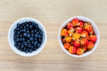 two white ceramic bowls with ripe berries: in one cloudberry, in the other blueberries on a wooden table