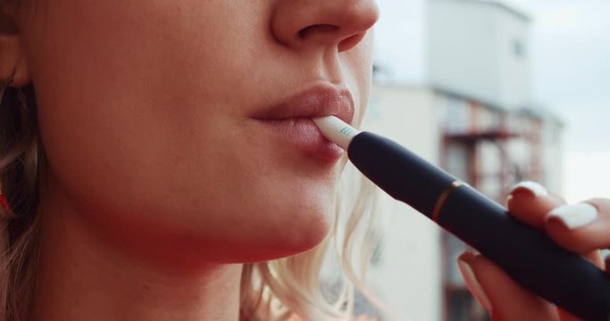 Close-up pretty woman with electronic cigarette outdoors. Young woman smoking tobacco stick heating and exhailing smoke.