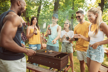 Group of happy friends having beer and barbecue party at sunny day. Resting together outdoor in a forest glade or backyard. Celebrating and relaxing, laughting. Summer lifestyle, friendship concept.