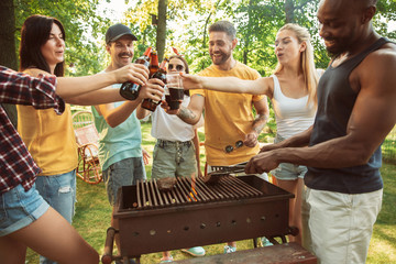 Group of happy friends having beer and barbecue party at sunny day. Resting together outdoor in a forest glade or backyard. Celebrating and relaxing, laughting. Summer lifestyle, friendship concept.