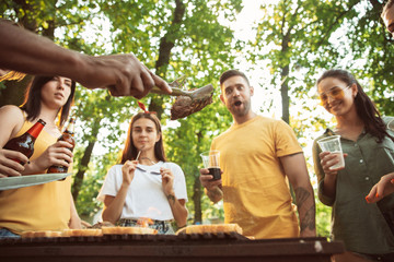 Group of happy friends having beer and barbecue party at sunny day. Resting together outdoor in a forest glade or backyard. Celebrating and relaxing, laughting. Summer lifestyle, friendship concept.