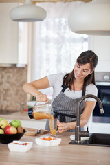 Beautiful smiling Caucasian woman in apron standing in kitchen and pouring fresh smoothie into glass.