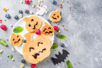 Halloween kids food treat. Funny pancakes - ghosts, jack o Lantern pumpkins, spider webs. Breakfast snack for child's Halloween party, with berries & candy,  gray stone table top view copy space