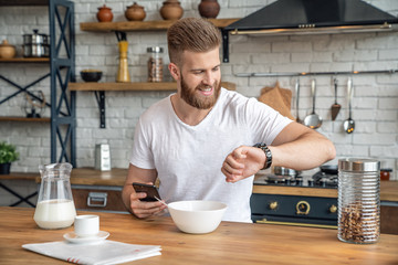 Handsome bearded man is sitting in the kitchen having breakfast cereals and milk, coffee and bananas. checking his mail on the phone and looking at his watch to make sure he is not late and smiling.