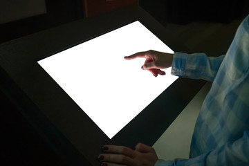 Mockup image - woman hand touching white empty interactive touchscreen display kiosk in dark room of modern technology museum. Mock up, template, scifi, education, futuristic and technology concept