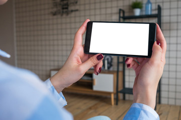 Over shoulder view: woman hands holding black smartphone with white blank screen in home interior. Mock up, copyspace, template, entertainment and technology concept