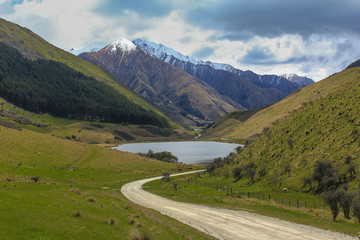 The view down a dirt track road to a small lake surronded by mountains and hills, snow sitting on top of the mountains.