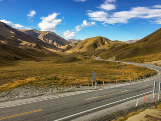 The stunning brown landscape of the rolling hills and mountains of the Lindis Pass, New Zealand with a road weaving through the landscape