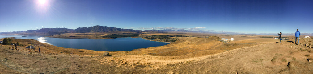 Fototapeta na wymiar Sweeping panoramic views of the spectacular golden Mackenzie Basin from the top of Mt John including Lake Tekapo with people in the foreground