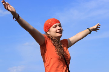 Young girl (woman) with long wavy brown hair, orange T-shirt (tee-shirt) and bandanna (scarf) is happy in summer sunny day and sky is blue with small clouds.