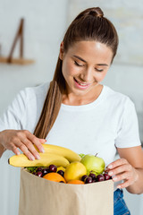 Obraz premium cheerful young woman looking at tasty fruits in paper bag