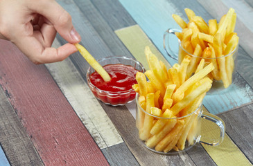 Hand dipping French fries in ketchup. French fries serve in two glass mugs on a wooden surface...
