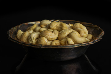 Lot of whole unsalted beige cashew in metal bowl isolated on black glass
