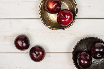 Group of six whole ripe red round plum in dark ceramic bowl in metal bowl flatlay on white wood
