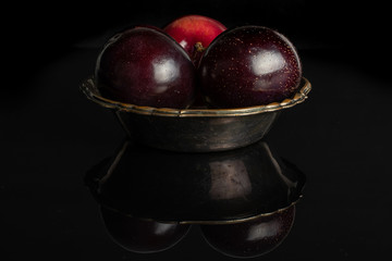 Group of three whole ripe red round plum in dark ceramic bowl isolated on black glass