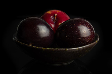 Group of three whole ripe red round plum in dark ceramic bowl isolated on black glass