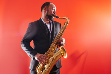 Portrait of professional musician saxophonist man in  suit plays jazz music on saxophone, red background in a photo studio - obrazy, fototapety, plakaty