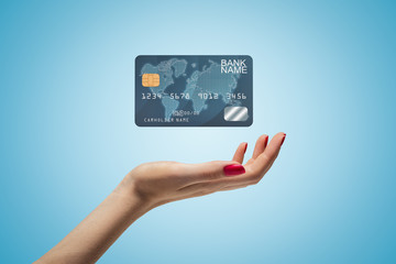 Side closeup of woman's hand facing up and levitating plastic bank card on light blue gradient background.