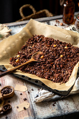 homemade baked chocolate granola with nuts, oat flakes on baking tray with parchment stands on grey wooden table