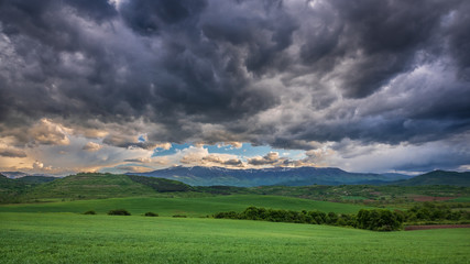 A dramatic landscape with green agriculture fields in the foreground, distant mountain range on the...