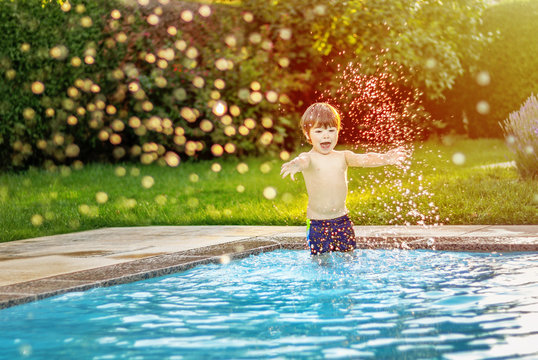 Happy little boy playing and havung fun in swimming pool splashing water drops in the air at sunset light. Summer lifestyle and leisure activity. Child enjoying summer holidays
