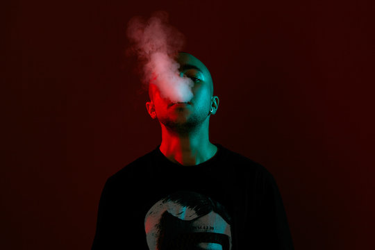 Close-up studio shot of a young bald guy vaping, blowing out a cloud of smoke on red background.