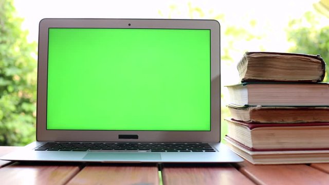 Closeup of modern laptop with green screen and lots old books on background. Technology development. Online education courses. Freelance lifestyle. Study app. Chroma key display. Sunlight.