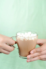 Clear glass with cocoa in the hand of a teenager. The drink is complemented by marshmallows. Hand with a glass on a light background. Close-up.