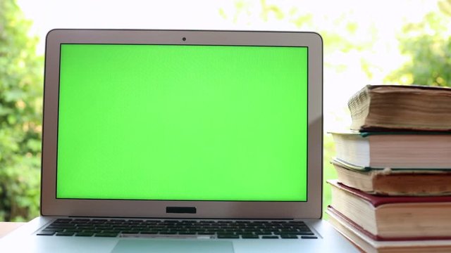 Closeup view of modern green screen laptop with old books on the table with sunny nature on blur background. Chromakey notebook monitor, mock-up for free application content. App developing, internet.