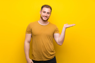 Blonde man over isolated yellow wall holding copyspace imaginary on the palm to insert an ad