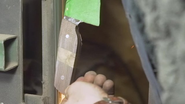 Extreme close-up shot of knifemaker making sparks while sanding the back of a kitchen knife's textured wood handle on a belt sander bench in his workshop - Slow Motion