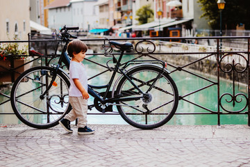 Fototapeta na wymiar Little boy walking over bridge of Thiou River in Annecy, France near parked bicycle. Child in the city. Little tourist, summer lifestyle. Discovering Europe, traveling with kid.