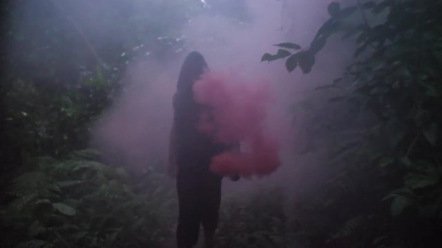 Dark figure walking on a narrow jungle patch waving red smoke. Man with martial arts moves as he walks through a tropical forest. Face concealed by darkness and smoke.