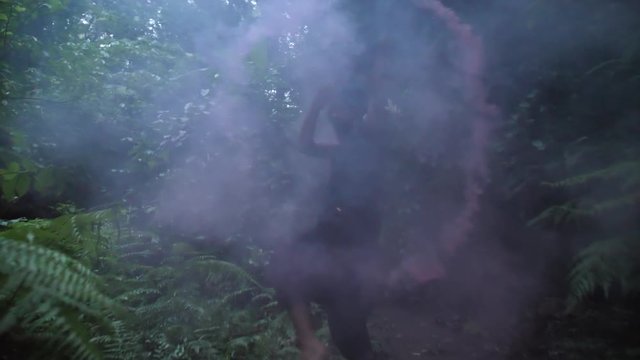 Spooky figure in shadow waving red smoke grenade in a tropical forest. Man in jungle with smoke.
