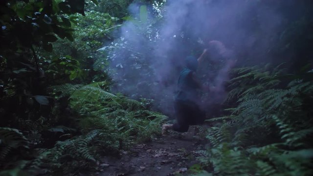 Slow motion view of man in jungle waving red smoke with martial arts movements. Dark, mysterious figure in tropical rainforest with face concealed by hood and smoke. PUSH IN.